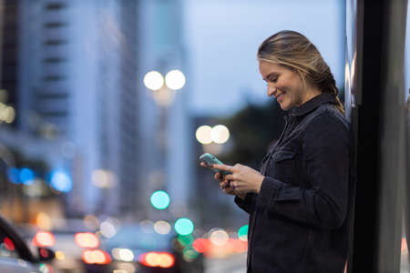 Young woman using a smartphone at night time with city view landscape in the background. High quality photo. photo. Mobile phone, technology, urban concept.の写真素材