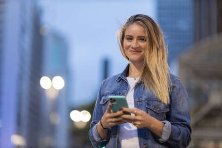 Young woman using a smartphone at night time with city view landscape in the background. High quality photo. photo. Mobile phone, technology, urban concept.の写真素材