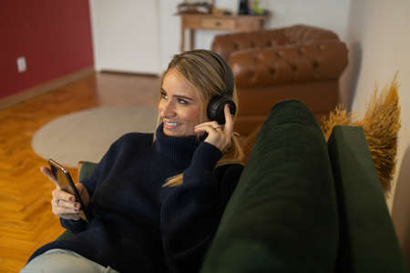 Young woman listening to music with headphones in her ears in her bed in her bedroom inside her apartment. High quality photo. Mobile phone, technology, music, urban living, feel good concept.の写真素材