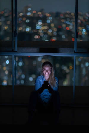 Young woman using a smartphone at night time with city view landscape in the background. High quality photo. photo. Mobile phone, technology, urban concept.の写真素材