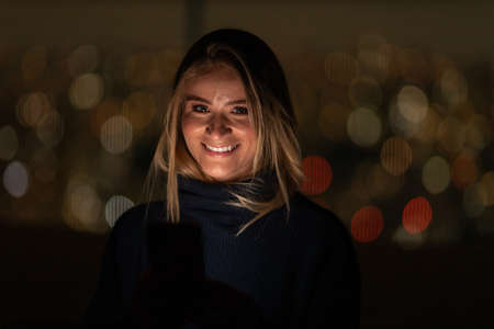 Young woman using a smartphone at night time with city view landscape in the background. High quality photo. photo. Mobile phone, technology, urban concept.の写真素材