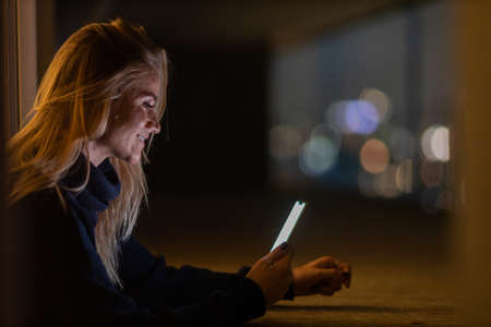 Young woman using a smartphone at night time with city view landscape in the background. High quality photo. photo. Mobile phone, technology, urban concept.の写真素材
