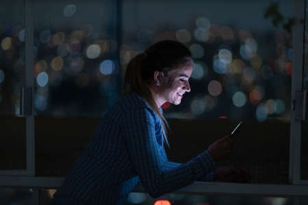 Young woman using a smartphone at night time with city view landscape in the background. High quality photo. photo. Mobile phone, technology, urban concept.の写真素材