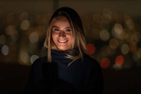 Young woman using a smartphone at night time with city view landscape in the background. High quality photo. photo. Mobile phone, technology, urban concept.の写真素材
