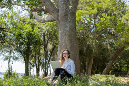 Young woman using a laptop at day time with a green park in the background. High quality photo. Mobile phone, technology, urban concept.の写真素材