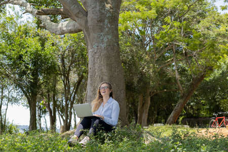 Young woman using a laptop at day time with a green park in the background. High quality photo. Mobile phone, technology, urban concept.の写真素材