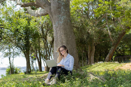 Young woman using a laptop at day time with a green park in the background. High quality photo. Mobile phone, technology, urban concept.の写真素材