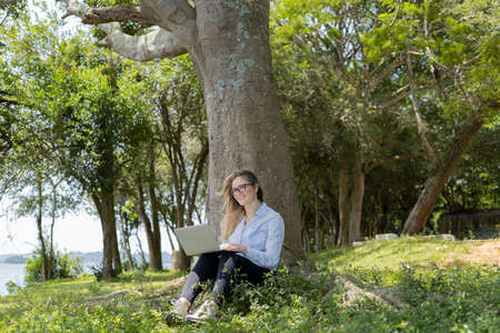 Young woman using a laptop at day time with a green park in the background. High quality photo. Mobile phone, technology, urban concept.の写真素材