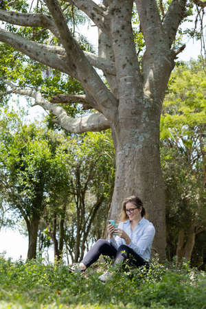 Young woman using a laptop at day time with a green park in the background. High quality photo. Mobile phone, technology, urban concept.の写真素材