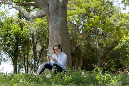 Portrait of smiling young woman at a park on a beautiful sunny day. Woman with open arms in front of a big a tree. Green and nature background. high quality photoの写真素材