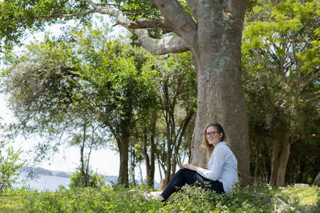 Portrait of smiling young woman at a park on a beautiful sunny day. Woman with open arms in front of a big a tree. Green and nature background. high quality photoの写真素材