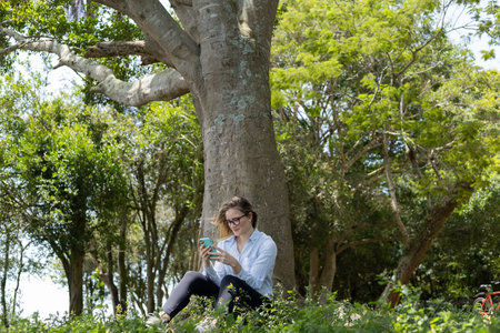 Portrait of smiling young woman at a park on a beautiful sunny day. Woman with open arms in front of a big a tree. Green and nature background. high quality photoの写真素材