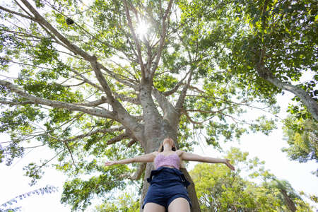 Portrait of smiling young woman at a park on a beautiful sunny day. Woman with open arms in front of a big a tree. Green and nature background. high quality photoの写真素材