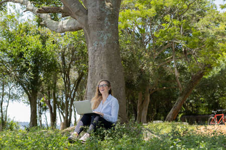 Portrait of smiling young woman at a park on a beautiful sunny day. Woman with open arms in front of a big a tree. Green and nature background. high quality photoの写真素材