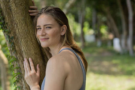Portrait of smiling young woman at a park on a beautiful sunny day. Woman with open arms in front of a big a tree. Green and nature background. high quality photoの写真素材