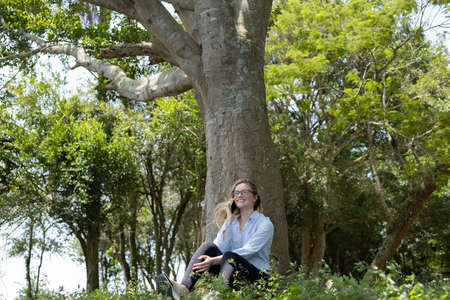 Portrait of smiling young woman at a park on a beautiful sunny day. Woman with open arms in front of a big a tree. Green and nature background. high quality photoの写真素材
