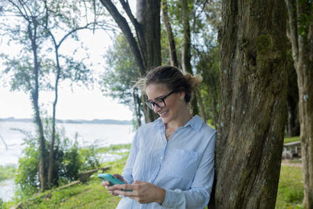 Portrait of smiling young woman at a park on a beautiful sunny day. Woman with open arms in front of a big a tree. Green and nature background. high quality photoの写真素材