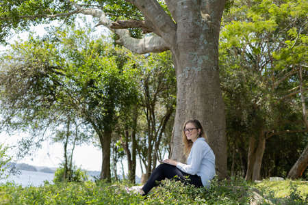 Portrait of smiling young woman at a park on a beautiful sunny day. Woman with open arms in front of a big a tree. Green and nature background. high quality photoの写真素材