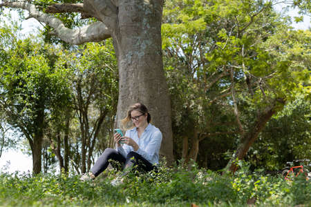 Portrait of smiling young woman at a park on a beautiful sunny day. Woman with open arms in front of a big a tree. Green and nature background. high quality photoの写真素材