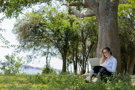 Young woman using a laptop at day time with a green park in the background. High quality photo. Mobile phone, technology, urban concept.の写真素材