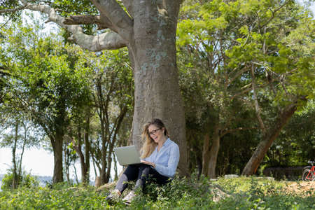 Young woman using a laptop at day time with a green park in the background. High quality photo. Mobile phone, technology, urban concept.の写真素材