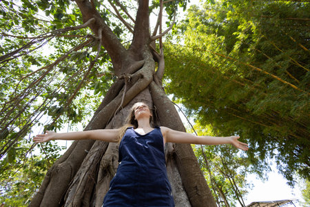 Portrait of smiling young woman at a park on a beautiful sunny day. Woman with open arms in front of a big a tree. Green and nature background. high quality photoの写真素材