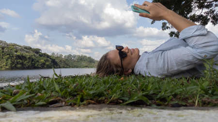 Young woman with sunglasses using a smartphone at day time at a green park with a lake - body of water. in the background High quality photo. Mobile phone, technology, urban concept. High quality photo Young woman with sunglasses lyind down on the grass using a smartphone at a green park with a lake - body of water. in the background High quality photo. Mobile phone, technology, urban concept.の写真素材