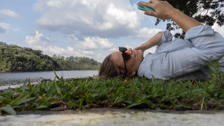 Young woman with sunglasses using a smartphone at day time at a green park with a lake - body of water. in the background High quality photo. Mobile phone, technology, urban concept. High quality photo Young woman with sunglasses lyind down on the grass using a smartphone at a green park with a lake - body of water. in the background High quality photo. Mobile phone, technology, urban concept.の写真素材