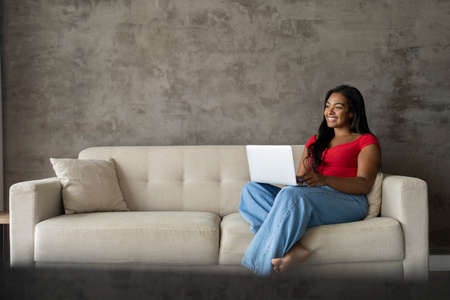 Young black woman working at home with laptop on her lap lying down on her couch in the living room. Home office concept. Gray notebook for working. high quality photoの写真素材