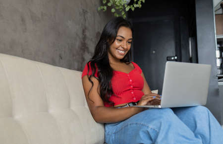 Young black woman working at home with laptop on her lap lying down on her couch in the living room. Home office concept. Gray notebook for working. high quality photoの写真素材