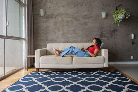 Young black woman working at home with laptop on her lap lying down on her couch in the living room. Home office concept. Gray notebook for working. high quality photoの写真素材