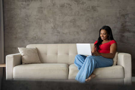 Young black woman working at home with laptop on her lap lying down on her couch in the living room. Home office concept. Gray notebook for working. high quality photoの写真素材