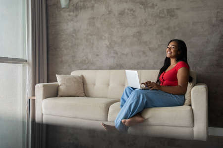 Young black woman working at home with laptop on her lap lying down on her couch in the living room. Home office concept. Gray notebook for working. high quality photoの写真素材