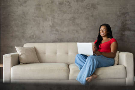 Young black woman working at home with laptop on her lap lying down on her couch in the living room. Home office concept. Gray notebook for working. high quality photoの写真素材