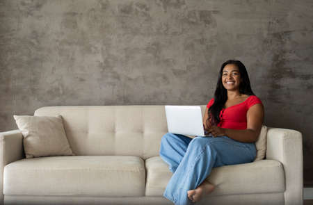 Young black woman working at home with laptop on her lap lying down on her couch in the living room. Home office concept. Gray notebook for working. high quality photoの写真素材