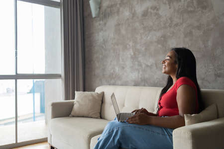 Young black woman working at home with laptop on her lap lying down on her couch in the living room. Home office concept. Gray notebook for working. high quality photoの写真素材