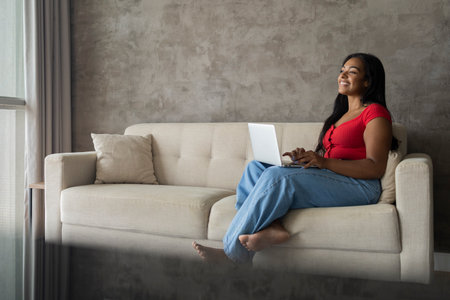 Young black woman working at home with laptop on her lap lying down on her couch in the living room. Home office concept. Gray notebook for working. high quality photoの写真素材