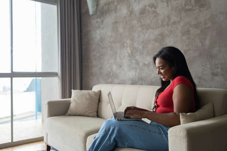 Young black woman working at home with laptop on her lap lying down on her couch in the living room. Home office concept. Gray notebook for working. high quality photoの写真素材