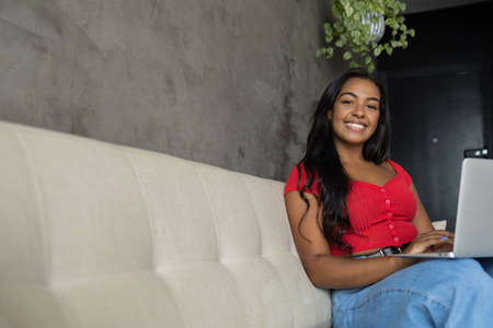Young black woman working at home with laptop on her lap lying down on her couch in the living room. Home office concept. Gray notebook for working. high quality photoの写真素材