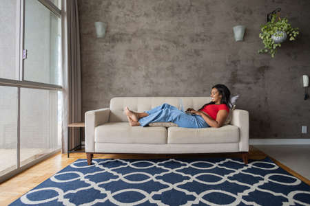 Young black woman working at home with laptop on her lap lying down on her couch in the living room. Home office concept. Gray notebook for working. high quality photoの写真素材