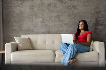 Young black woman working at home with laptop on her lap lying down on her couch in the living room. Home office concept. Gray notebook for working. high quality photoの写真素材