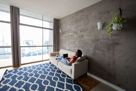 Young black woman working at home with laptop on her lap lying down on her couch in the living room. Home office concept. Gray notebook for working. high quality photoの写真素材