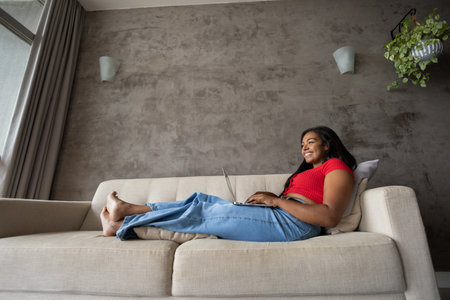 Young black woman working at home with laptop on her lap lying down on her couch in the living room. Home office concept. Gray notebook for working. high quality photoの写真素材