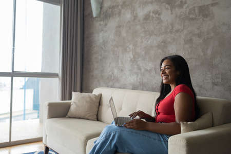Young black woman working at home with laptop on her lap lying down on her couch in the living room. Home office concept. Gray notebook for working. high quality photoの写真素材