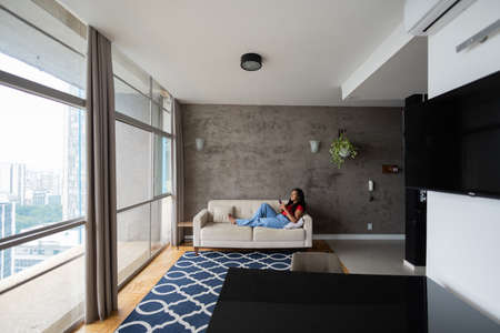 Young black woman working at home with laptop on her lap lying down on her couch in the living room. Home office concept. Gray notebook for working. high quality photoの写真素材