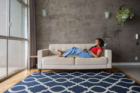 Young black woman working at home with laptop on her lap lying down on her couch in the living room. Home office concept. Gray notebook for working. high quality photoの写真素材