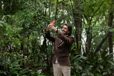 View of a young man with smartphone at daytime in a green park in the city. Taking a selfie - video call with smartphone. High quality photo.の写真素材