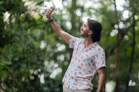 View of a young man with smartphone at daytime in a green park in the city. Taking a selfie - video call with smartphone. High quality photo.の写真素材