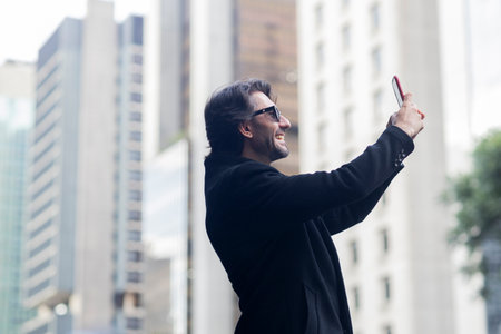 View of a well dressed young man with sunglasses and smartphone at daytime with city view landscape in the background. Taking a selfie - video call with smartphone. High quality photo.の写真素材