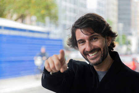 View of a young man pointing and looking at camera at daytime with city landscape in the background....